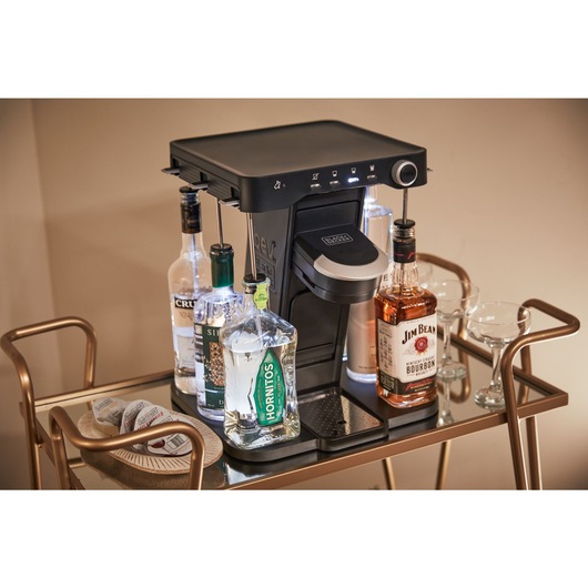 bev by BLACK+DECKER™ cocktail maker on a gold bar cart in a living room with assorted capsules and glassware