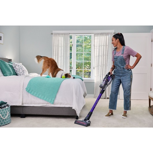 woman vacuuming bedroom carpet with BLACK+DECKER PowerseriesExtreme pet stick vacuum while dog is playing on the bed