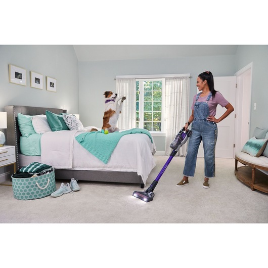 woman vacuuming bedroom carpet with BLACK+DECKER PowerseriesExtreme pet stick vacuum while dog is doing tricks on the bed