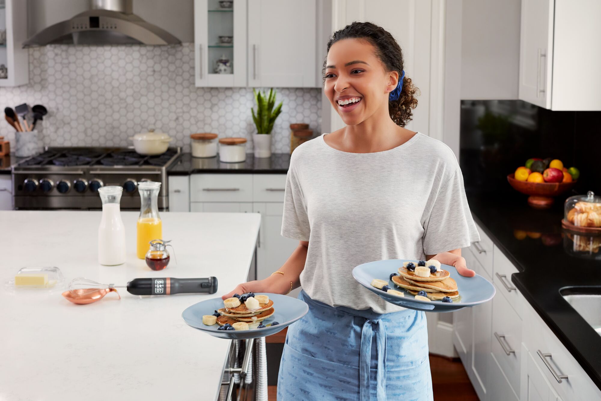 Talent holding two plates of pancakes with grey kitchen wand with whisk attachment visible on countertop in the background